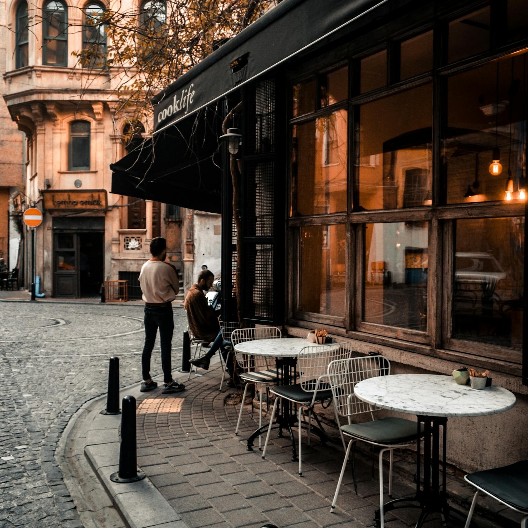 Facade of cafe with large windows and tables on sidewalk along road and faceless people resting in city
